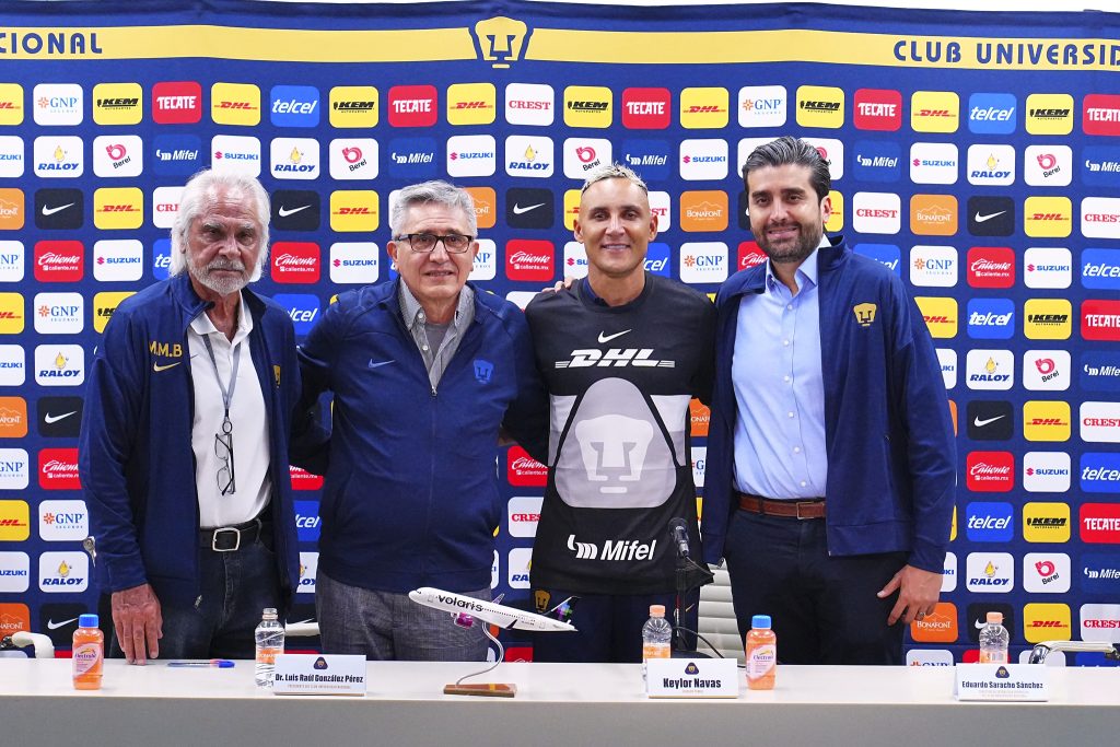 Keylor Navas posando con directivos de los Pumas durante su presentación oficial como nuevo portero del club, con un fondo de banners del equipo.