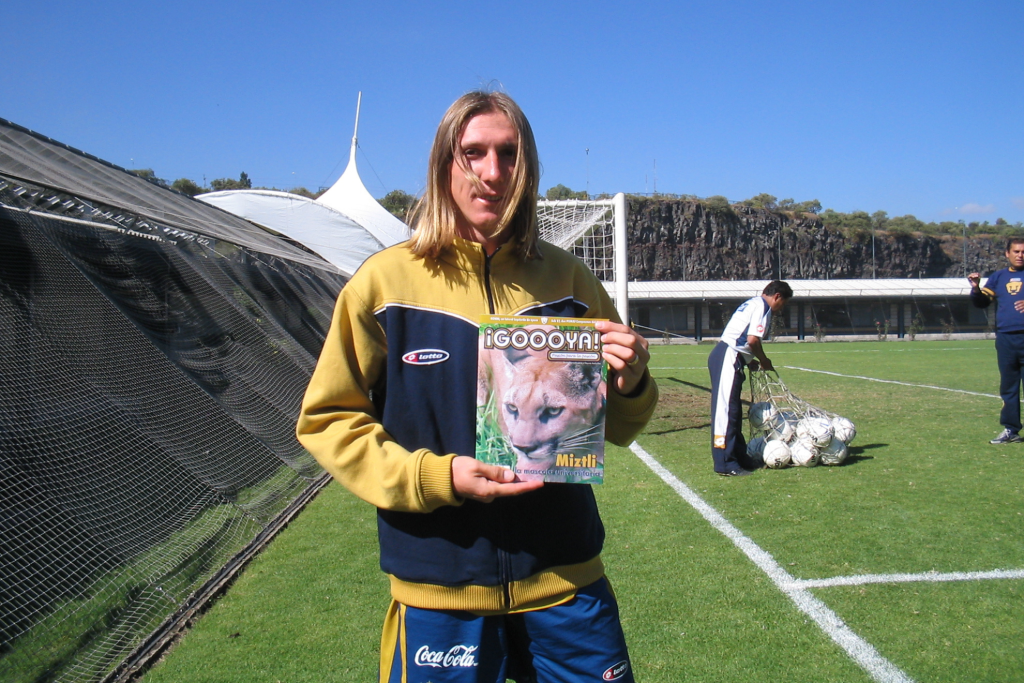 Leandro Augusto sosteniendo una revista ¡GOOOYA! en un campo de futbol, con un fondo de redes y una estructura al aire libre.