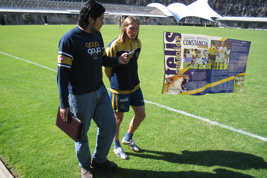Leandro Augusto, futbolista brasileño del Club Universidad Nacional, siendo entrevistado en un campo de entrenamiento, con un folleto en la mano que muestra una portada de revista.