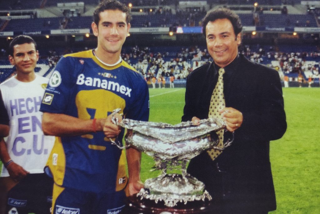 Un jugador de Pumas sostiene un trofeo en el estadio Santiago Bernabéu, acompañado por otro aficionado y un hombre con un traje. La celebración resalta la victoria del equipo mexicano sobre el Real Madrid.