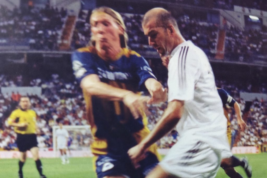 Un momento del partido entre Pumas y Real Madrid, donde un jugador de Pumas está en acción frente a Zinedine Zidane, con la multitud de fondo en el estadio Santiago Bernabéu.