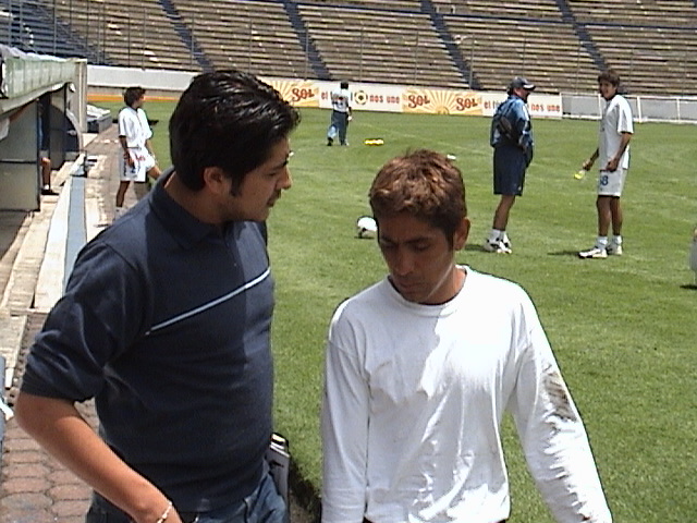 Dos hombres conversando en el campo de fútbol, con jugadores entrenando de fondo.