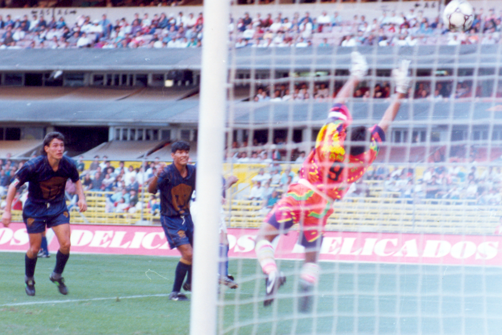 Jorge Campos, legendario portero mexicano, atajando un tiro en un partido de fútbol en el Estadio Olímpico Universitario, vistiendo su icónica camiseta multicolor.