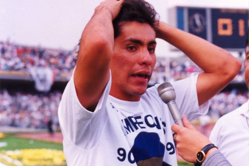 Jorge Campos, exfutbolista mexicano, siendo entrevistado en un evento deportivo mientras se lleva las manos a la cabeza, vistiendo una camiseta blanca con un diseño en el pecho. Fondos con público y un ambiente festivo.