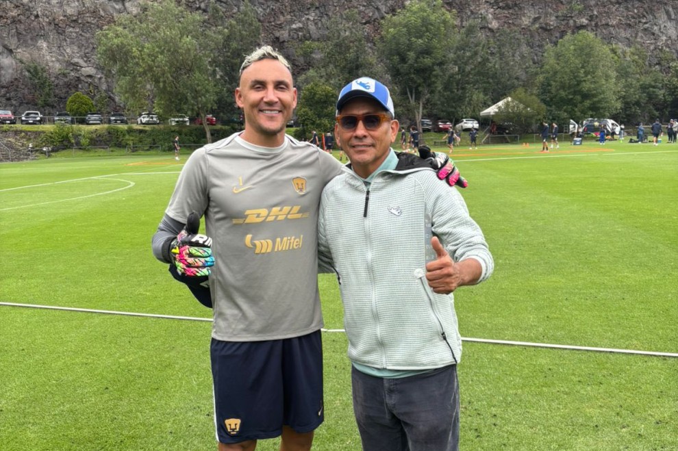 Jorge Campos, exfutbolista y portero de Pumas, posando junto a un actual portero en un campo de entrenamiento, ambos sonriendo y mostrando un gesto de pulgar hacia arriba.
